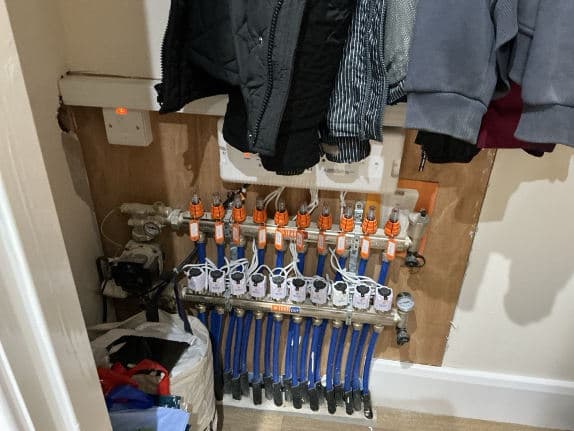 An underfloor heating manifold in a cupboard with coats hanging above it